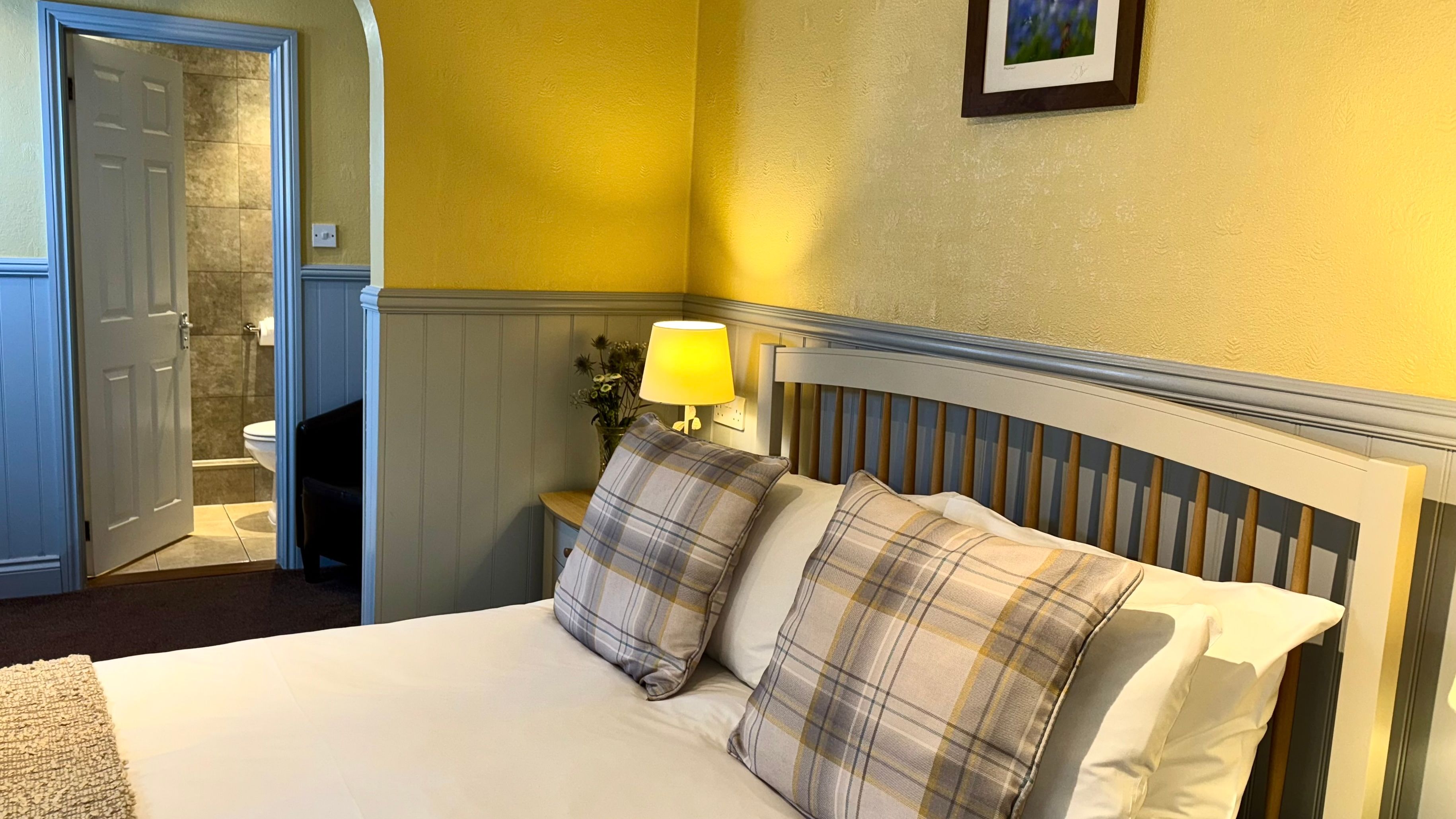 Cozy bedroom with a double bed, plaid pillows, bedside lamp, and open door to a tiled bathroom.