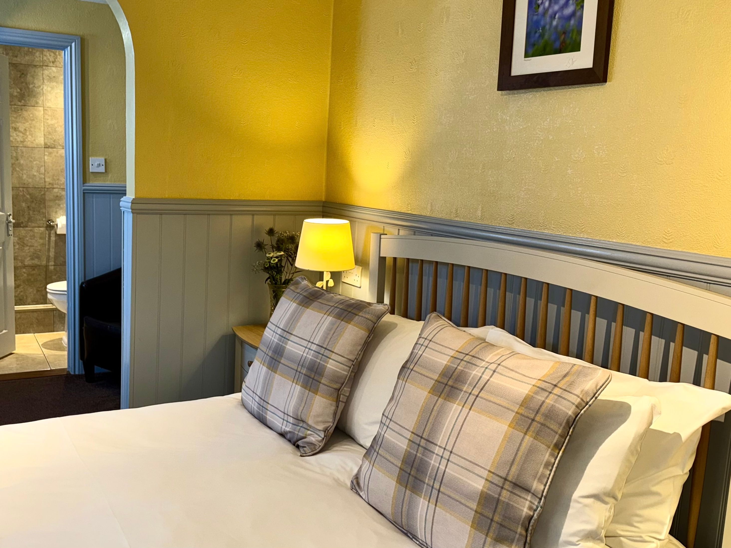 Cozy bedroom with a double bed, plaid pillows, bedside lamp, and open door to a tiled bathroom.