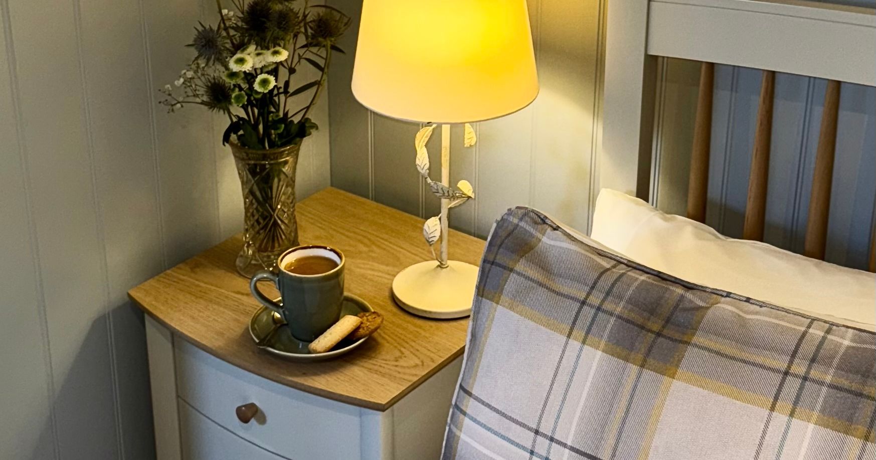 Cozy bedroom corner with a grey bedside table, cup of coffee, lamp, and plaid pillow