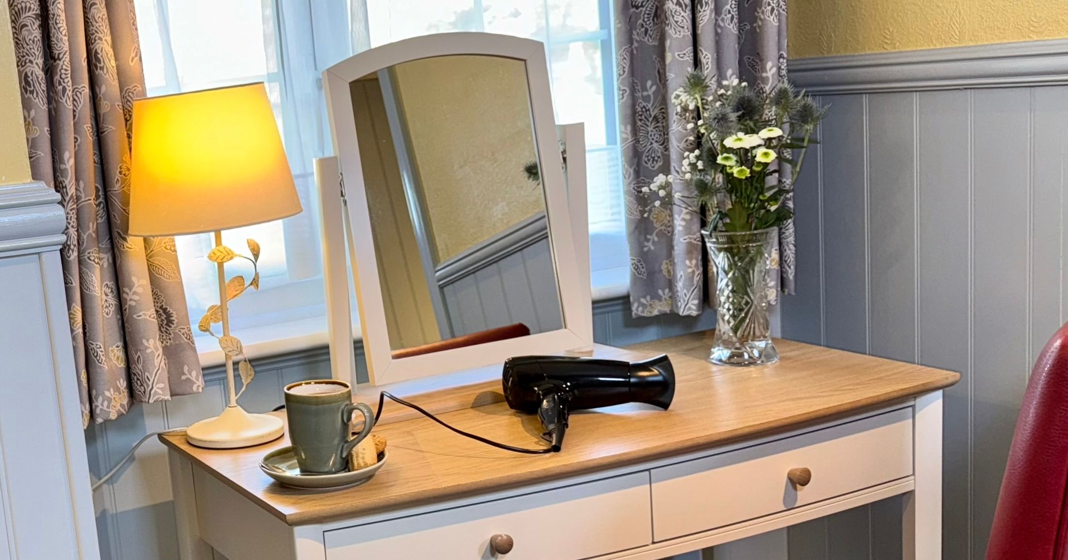 A vanity table with a mirror, a lamp, a vase of flowers, a cup and saucer, and a hair dryer.