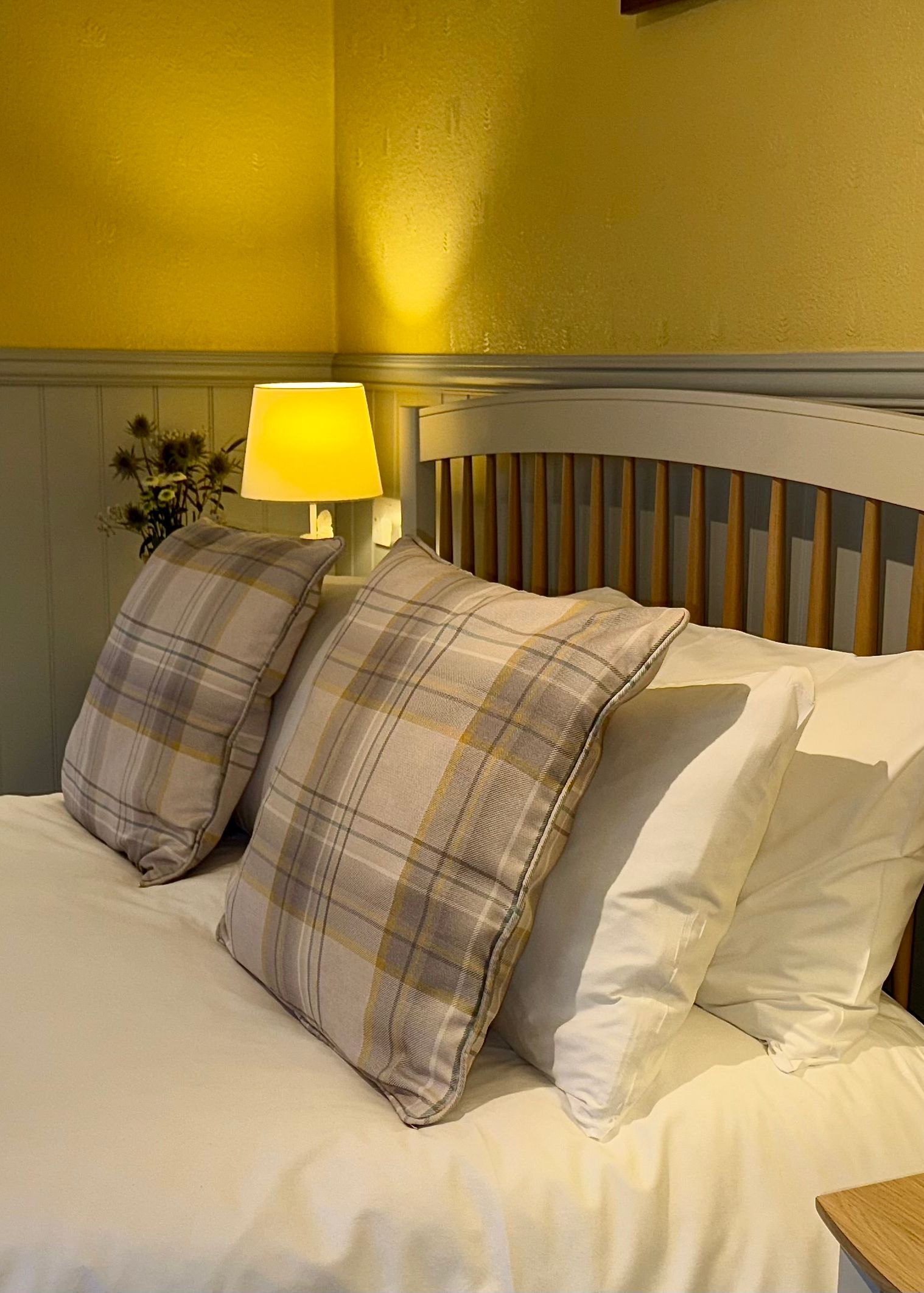 Cozy bedroom with a double bed featuring plaid pillows, two bedside lamps, and a view into a tiled bathroom.