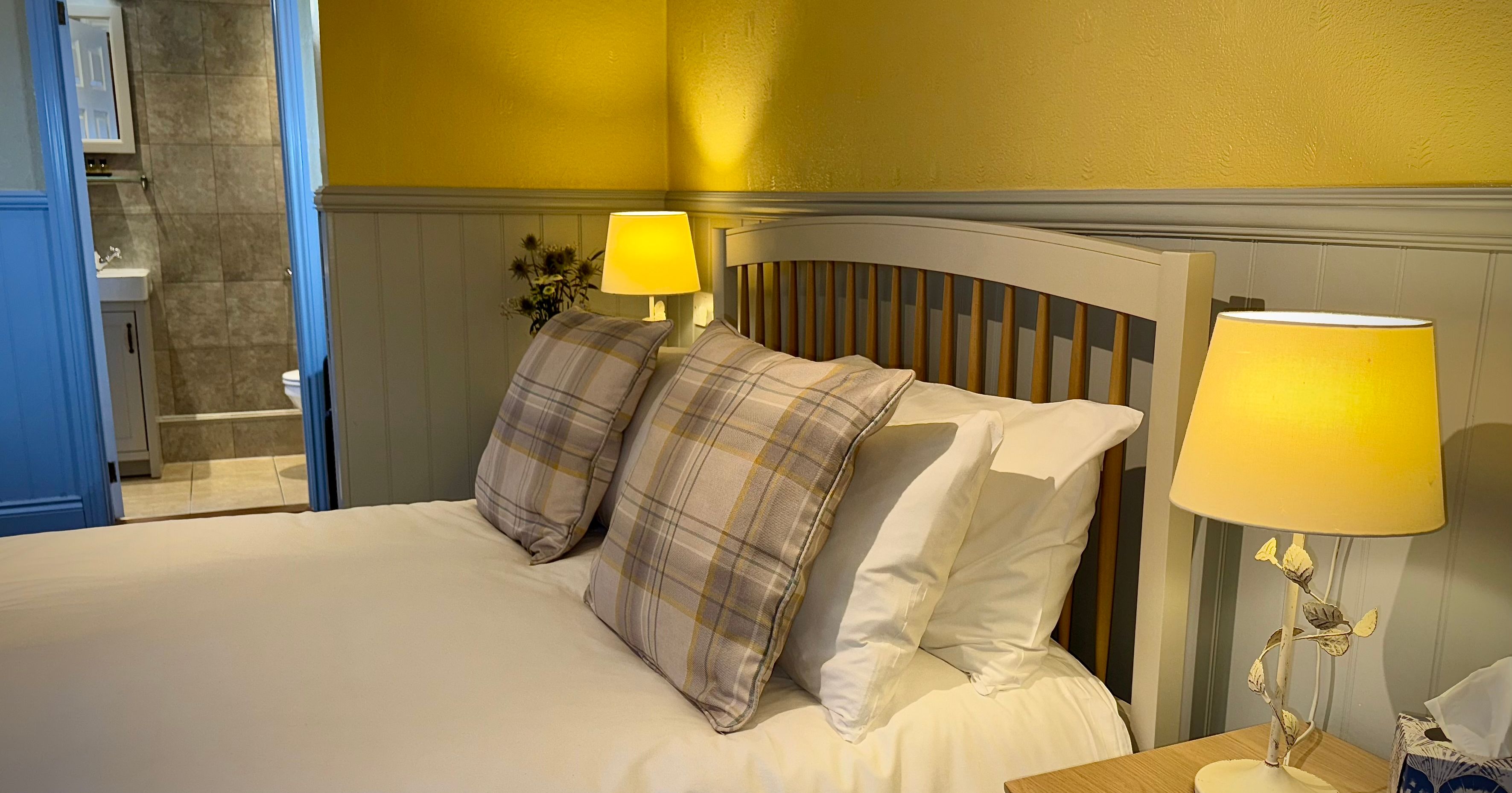Cozy bedroom with a double bed featuring plaid pillows, two bedside lamps, and a view into a tiled bathroom.