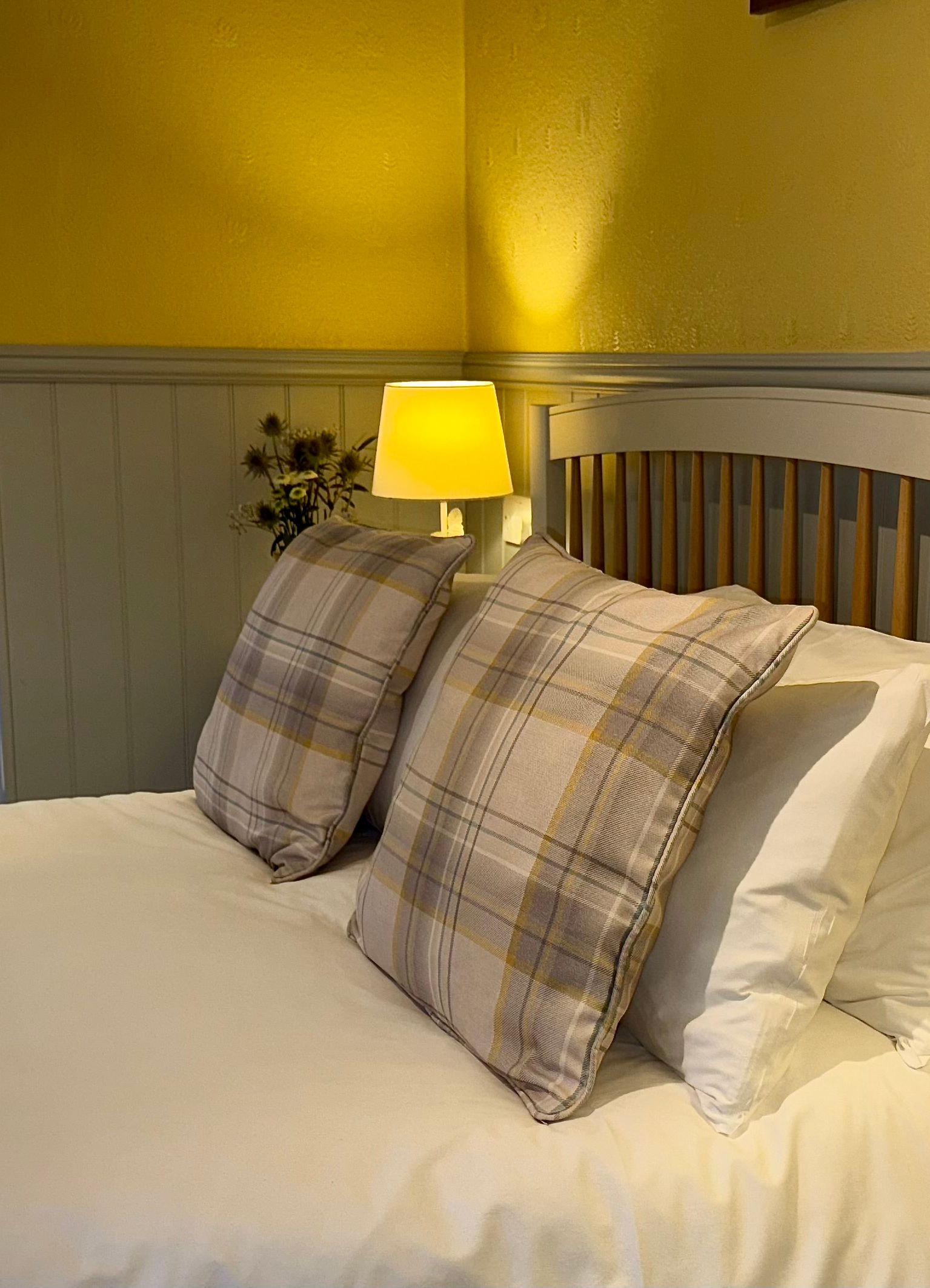 Cozy bedroom with a double bed featuring plaid pillows, two bedside lamps, and a view into a tiled bathroom.