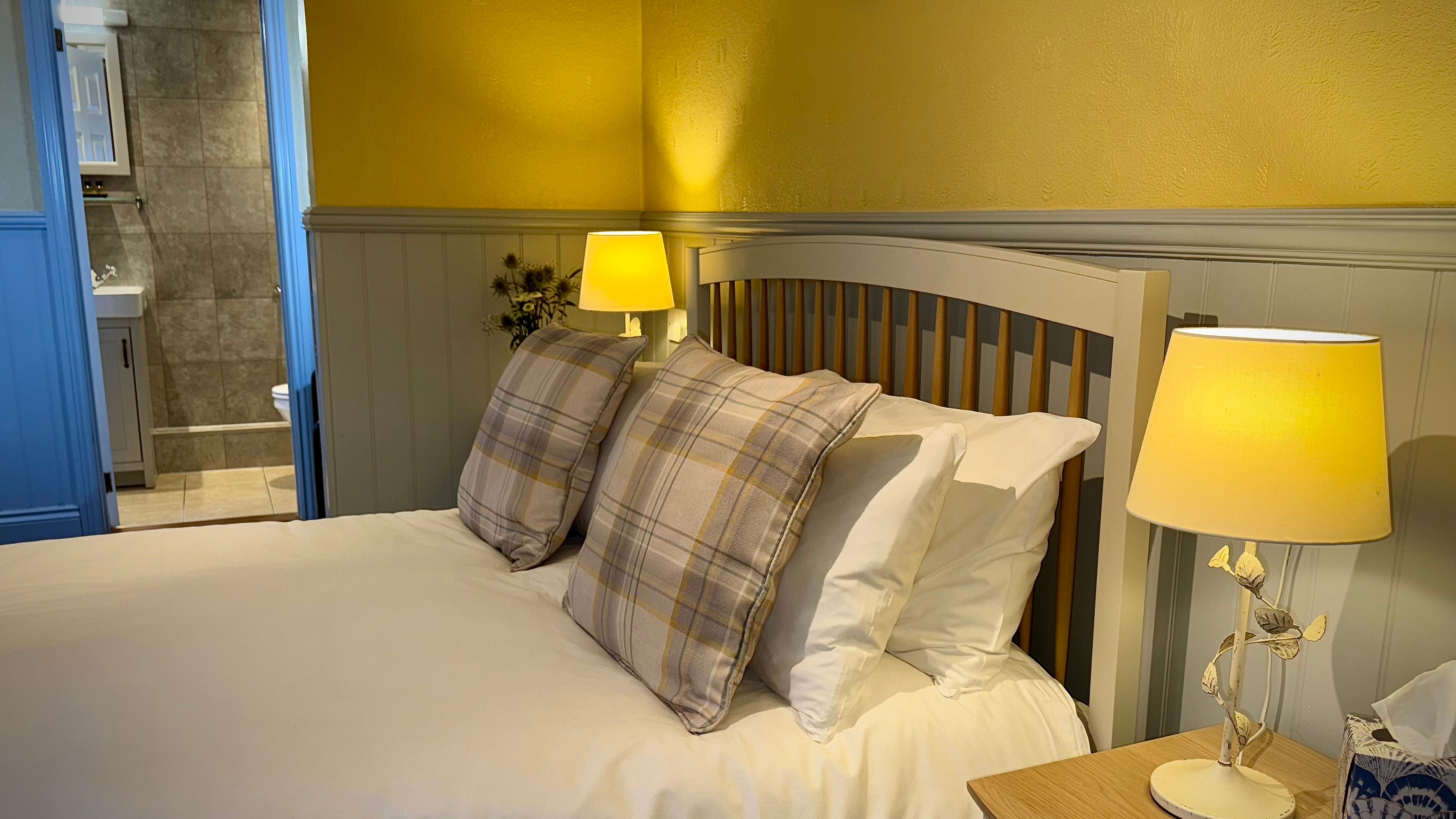 Cozy bedroom with a double bed featuring plaid pillows, two bedside lamps, and a view into a tiled bathroom.