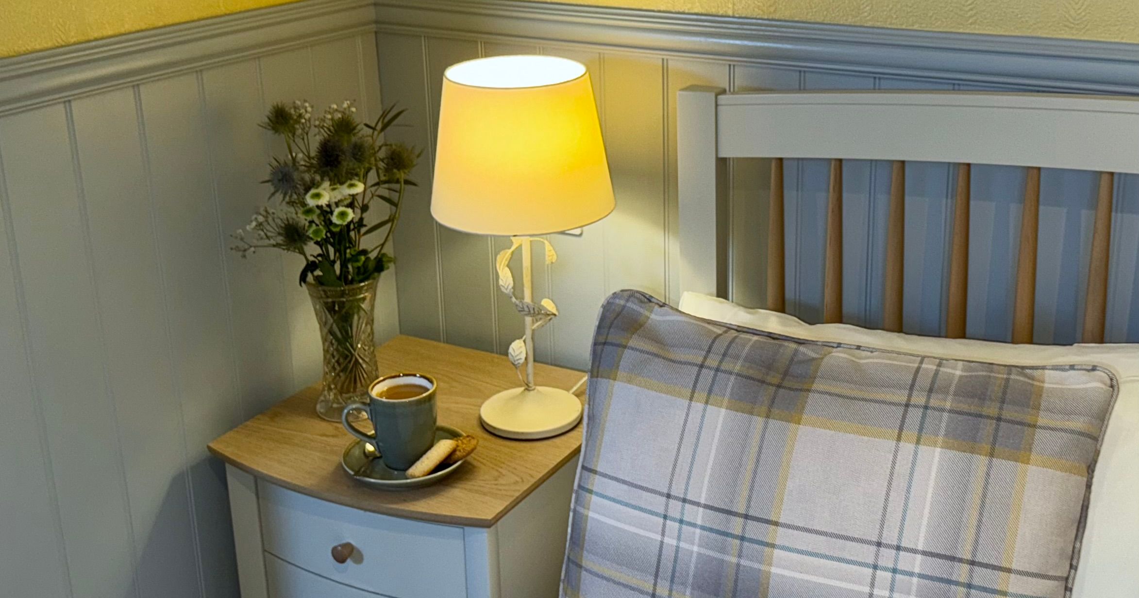 Cozy bedroom nook with lamp, cup of coffee, flowers, and plaid pillow