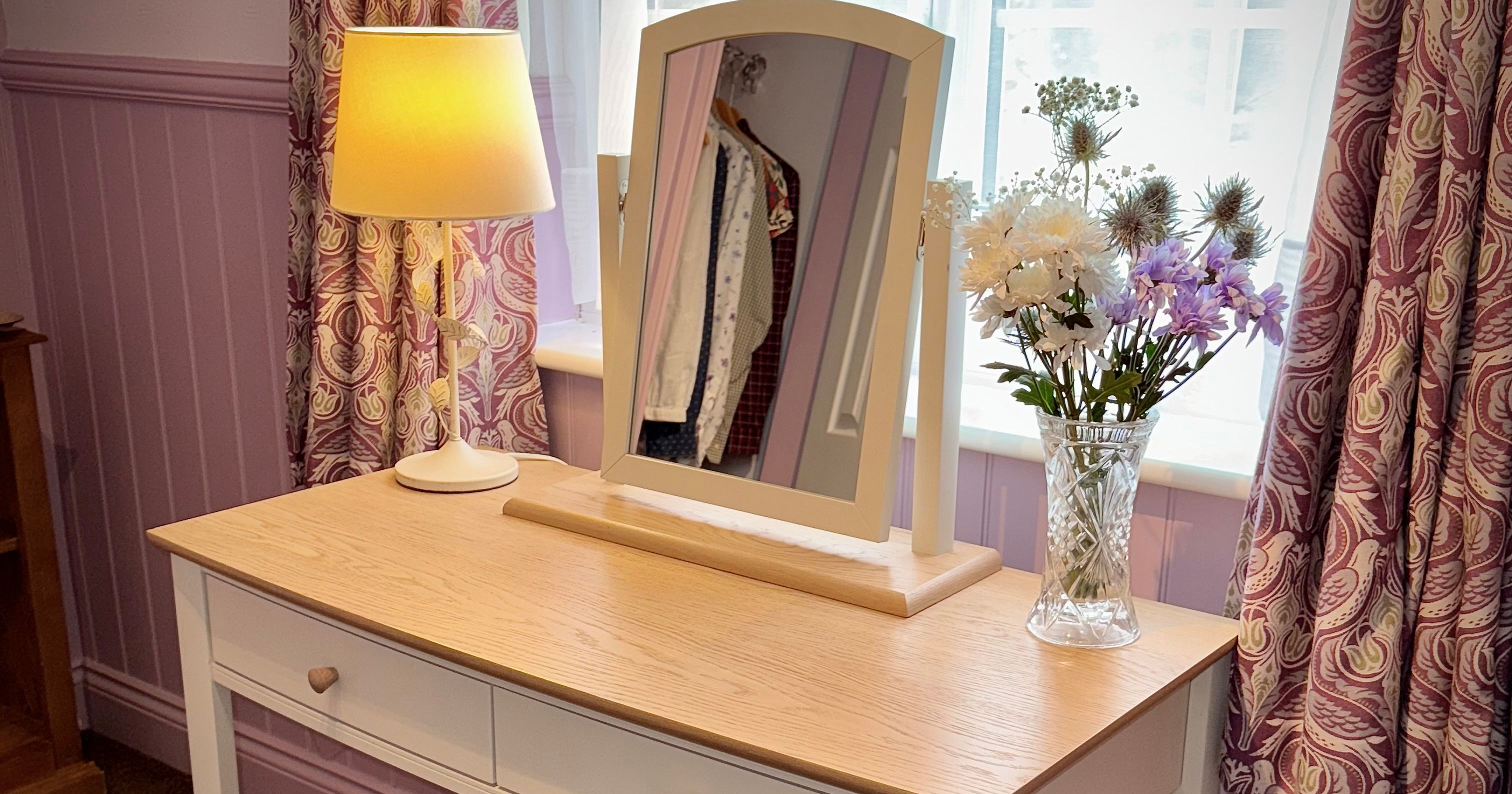 A wooden dressing table with a lamp, mirror, and vase of flowers by a window.