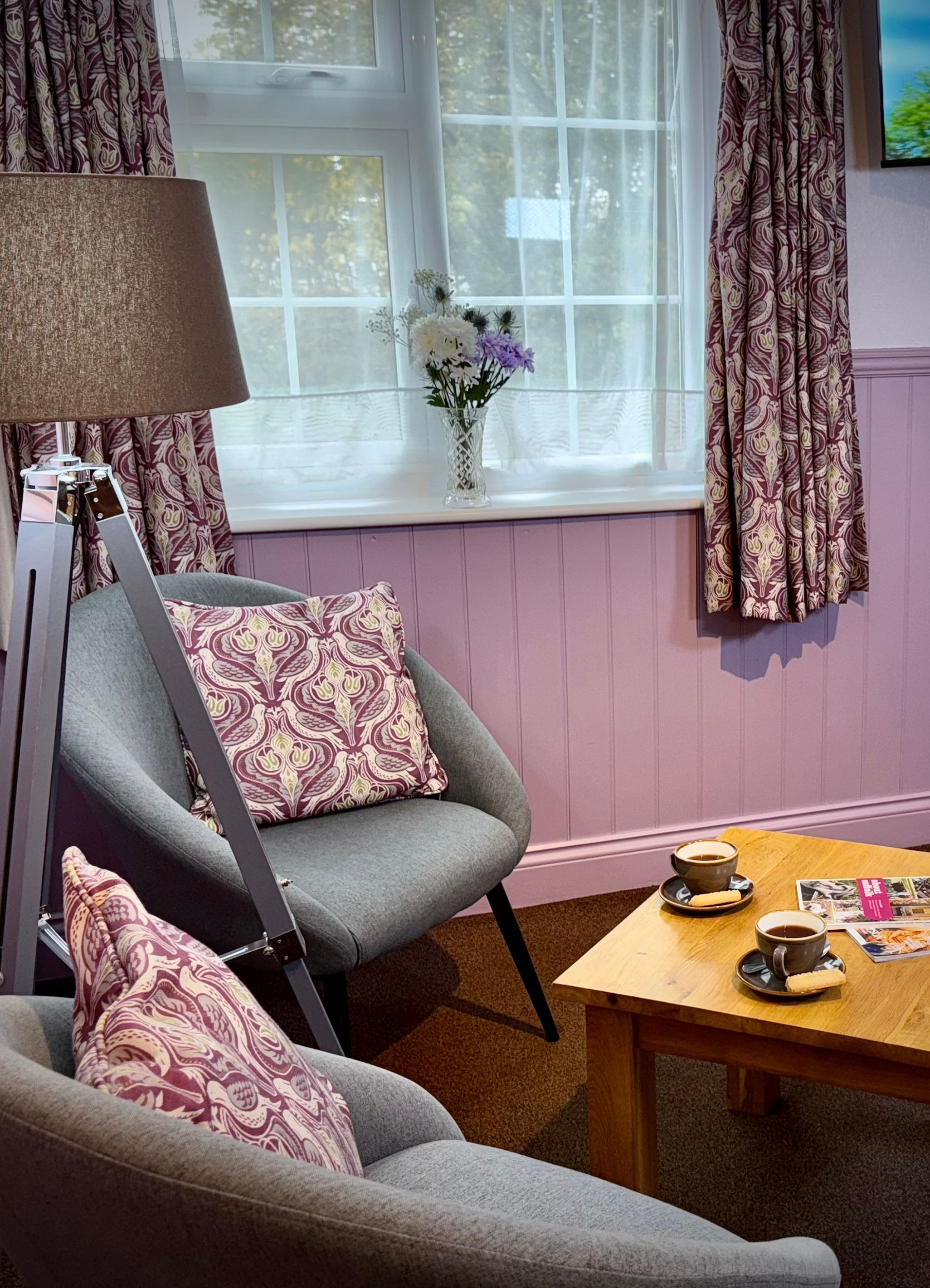 Cozy sitting area with two gray chairs, a wooden coffee table with cups and magazines, a lamp, and a window with patterned curtains.