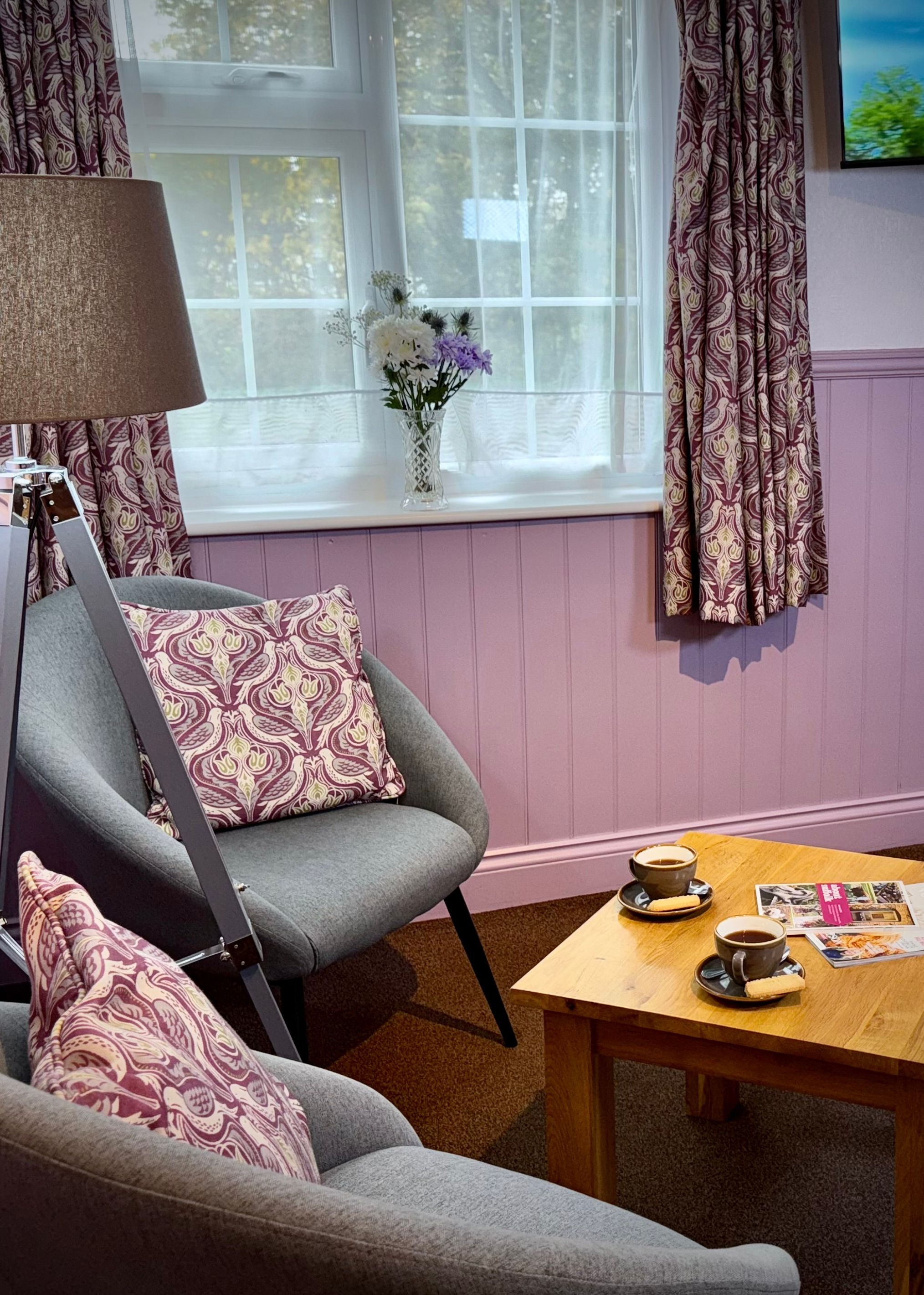 Cozy sitting area with two gray chairs, a wooden coffee table with cups and magazines, a lamp, and a window with patterned curtains.