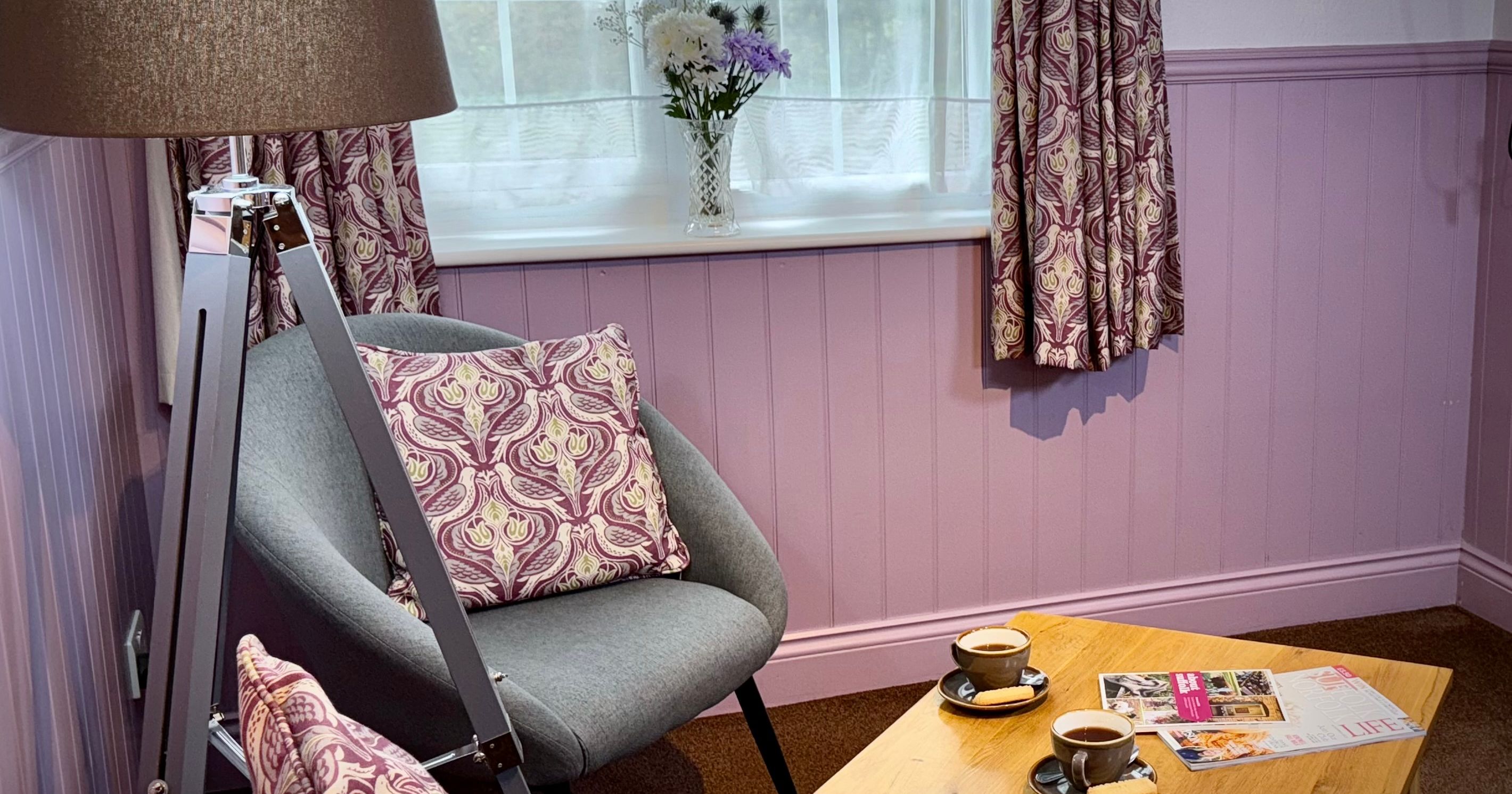 Cozy sitting area with two gray chairs, a wooden coffee table with cups and magazines, a lamp, and a window with patterned curtains.