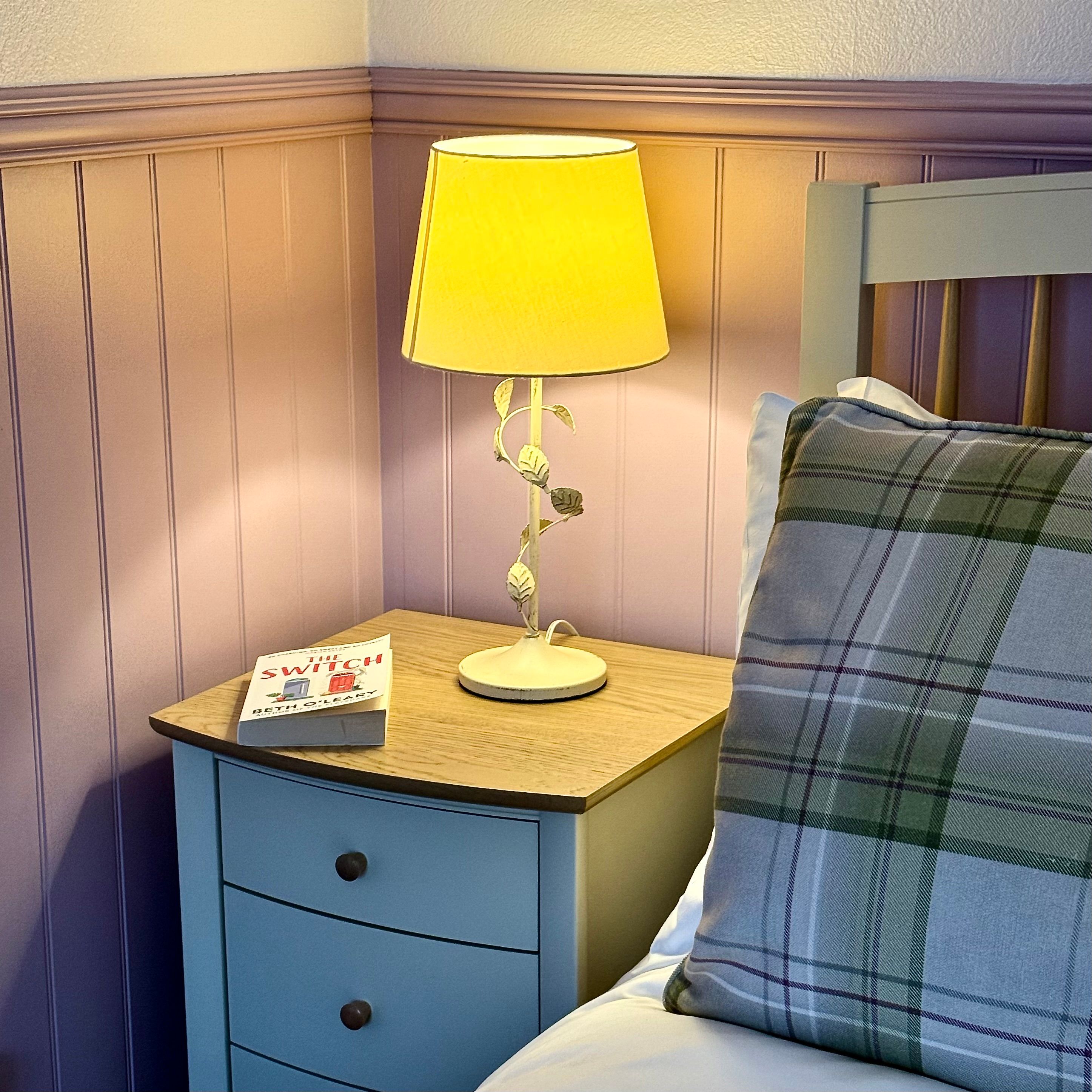 A cozy bedside table with a lamp, a book, and a plaid pillow on a bed.