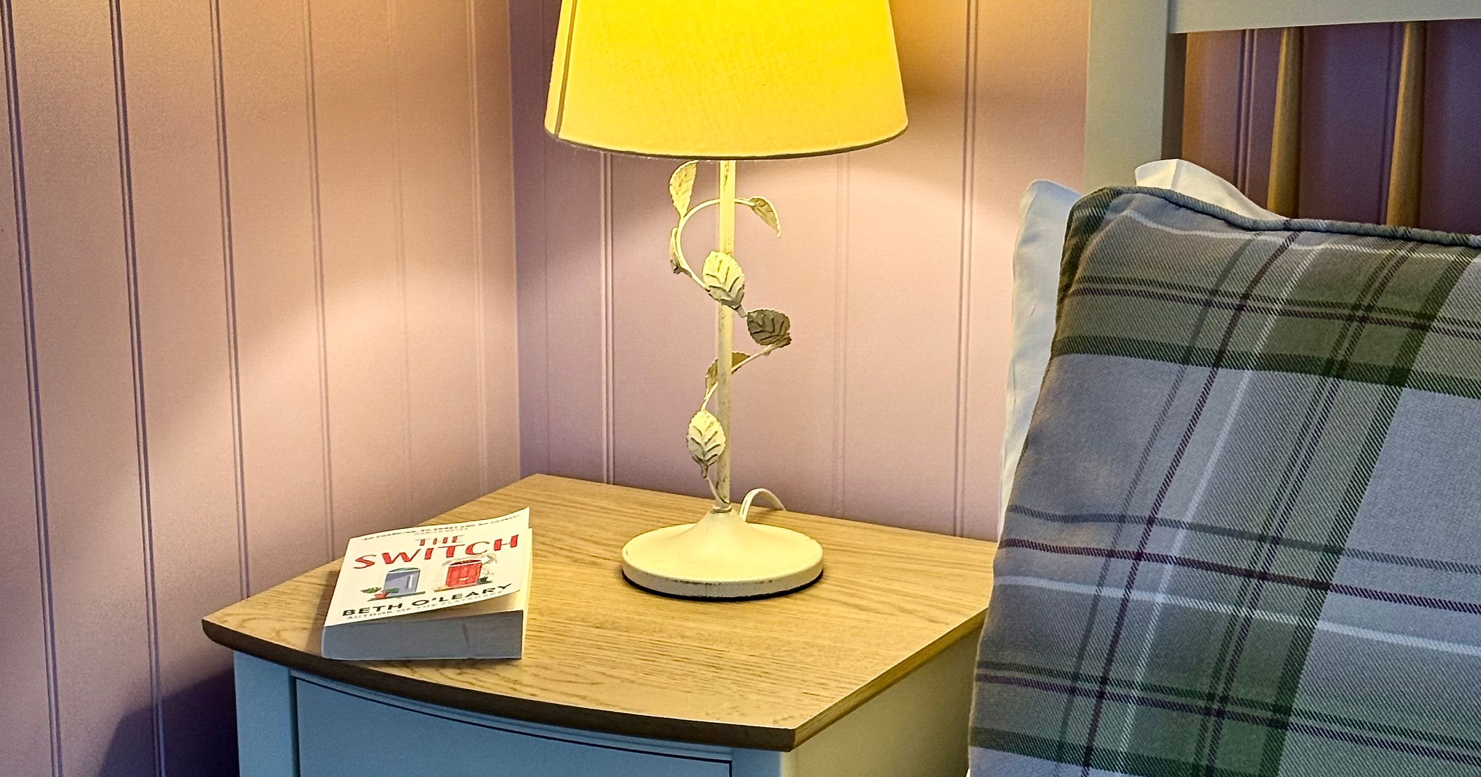 A cozy bedside table with a lamp, a book, and a plaid pillow on a bed.