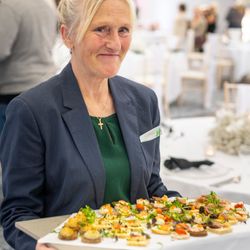A woman in a blazer and green shirt holding a tray of assorted appetizers at a formal event or banquet, with tables and people blurred in the background.