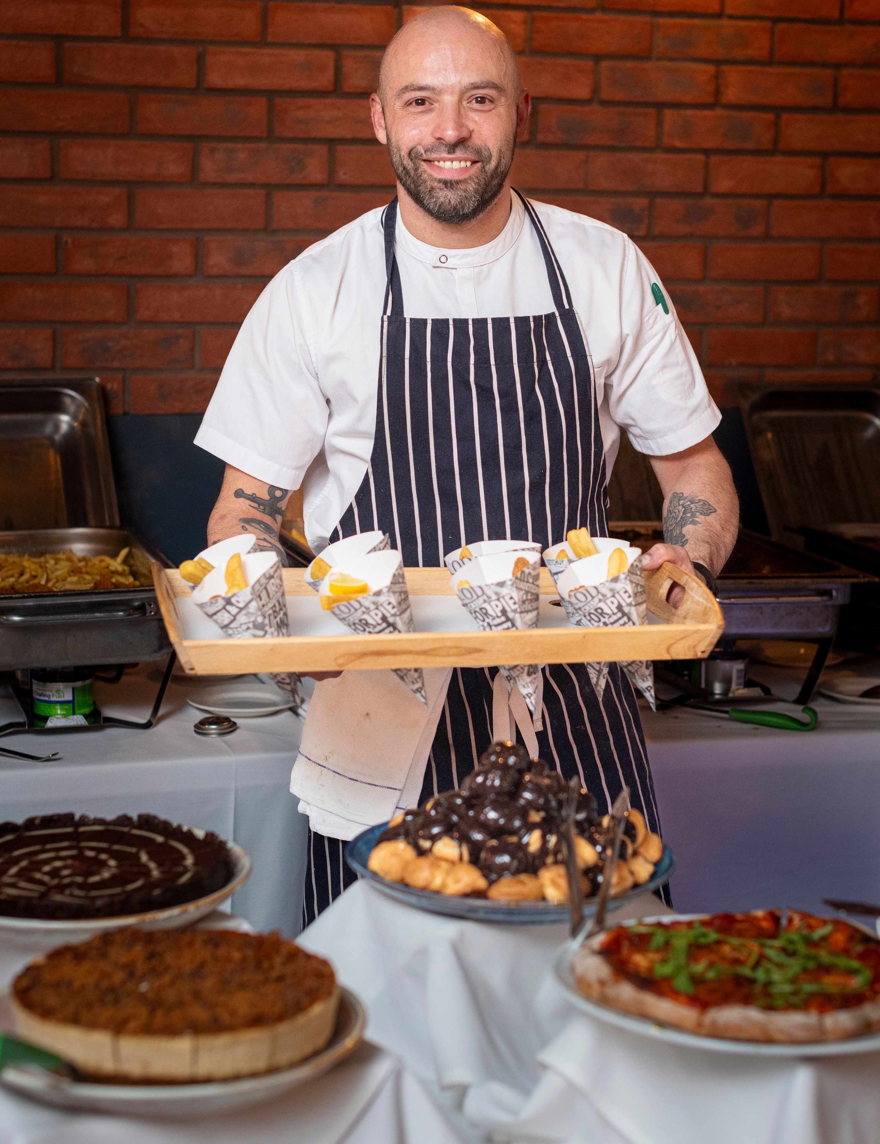 A smiling chef in a striped apron holding a tray with cones of French fries, standing behind a table with various desserts and dishes.