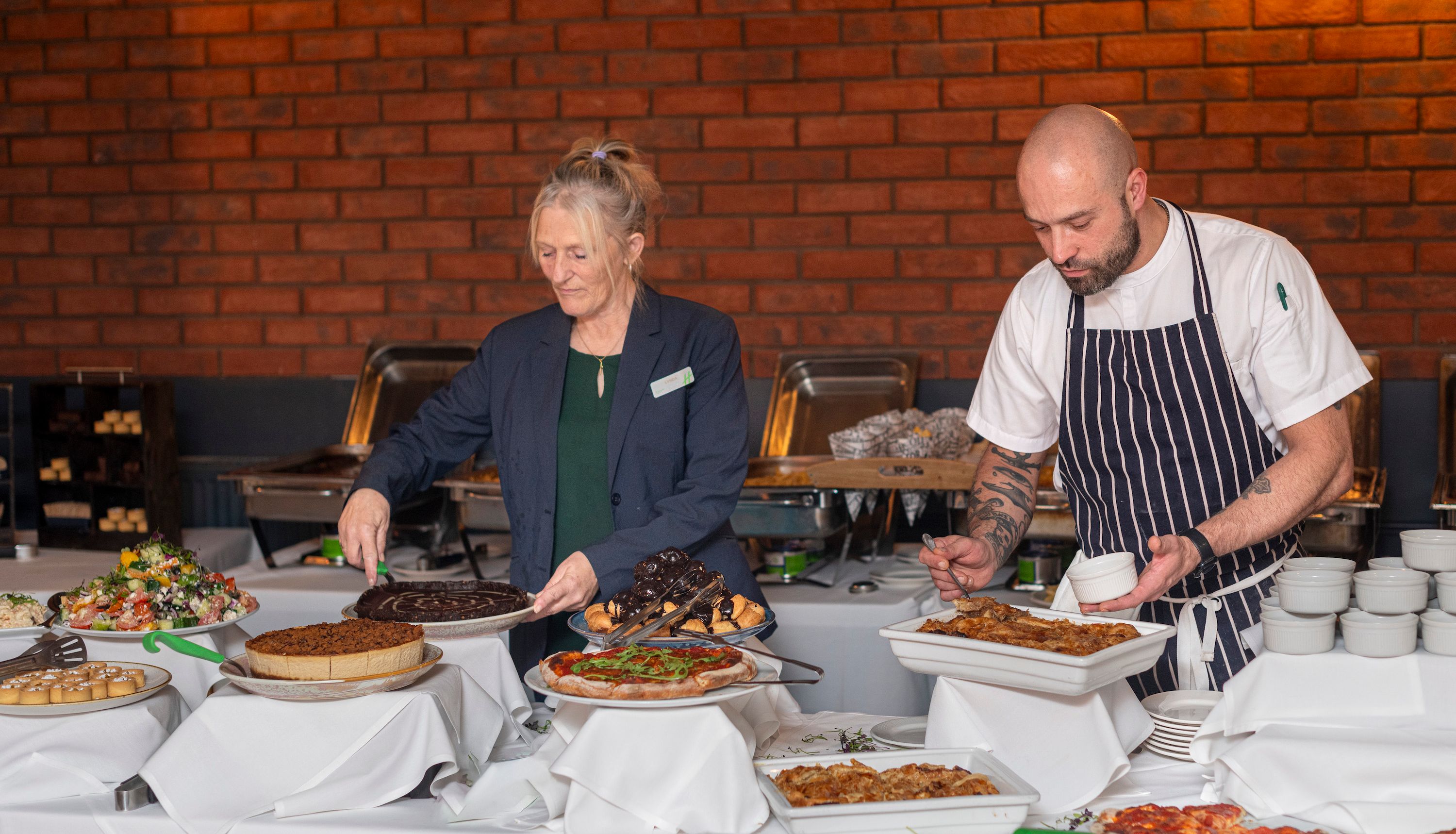 Two people serving food at a buffet table with various dishes, including salad, chocolate cake, tart, pizza, and pastries, in a restaurant setting with a brick wall background.