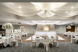 Elegant wedding reception hall decorated in white with round tables, floral centerpieces, draped ceiling fabric, a large 'LOVE' sign, and a long head table with a floral backdrop.
