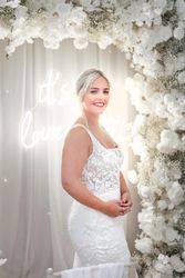 Bride in a white lace wedding dress standing in front of a floral arch decorated with white flowers and a neon sign in the background.