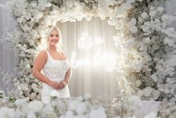 Smiling bride in a white lace wedding dress standing in front of a floral arch made of white flowers, with a neon sign in the background that reads 'it's a love story.'