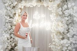 Bride in a white wedding dress holding a glass of champagne, standing under an arch made of white flowers with a neon sign that says 'it's a love story.'
