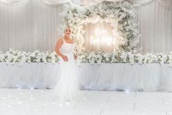 Bride in a white wedding dress standing in front of an elegant floral backdrop with a neon sign that says 'it's a love story,' surrounded by white flowers and drapery.
