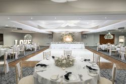 Elegant wedding reception hall decorated in white with round tables, floral centerpieces, a long head table, and illuminated 'LOVE' sign in the background.