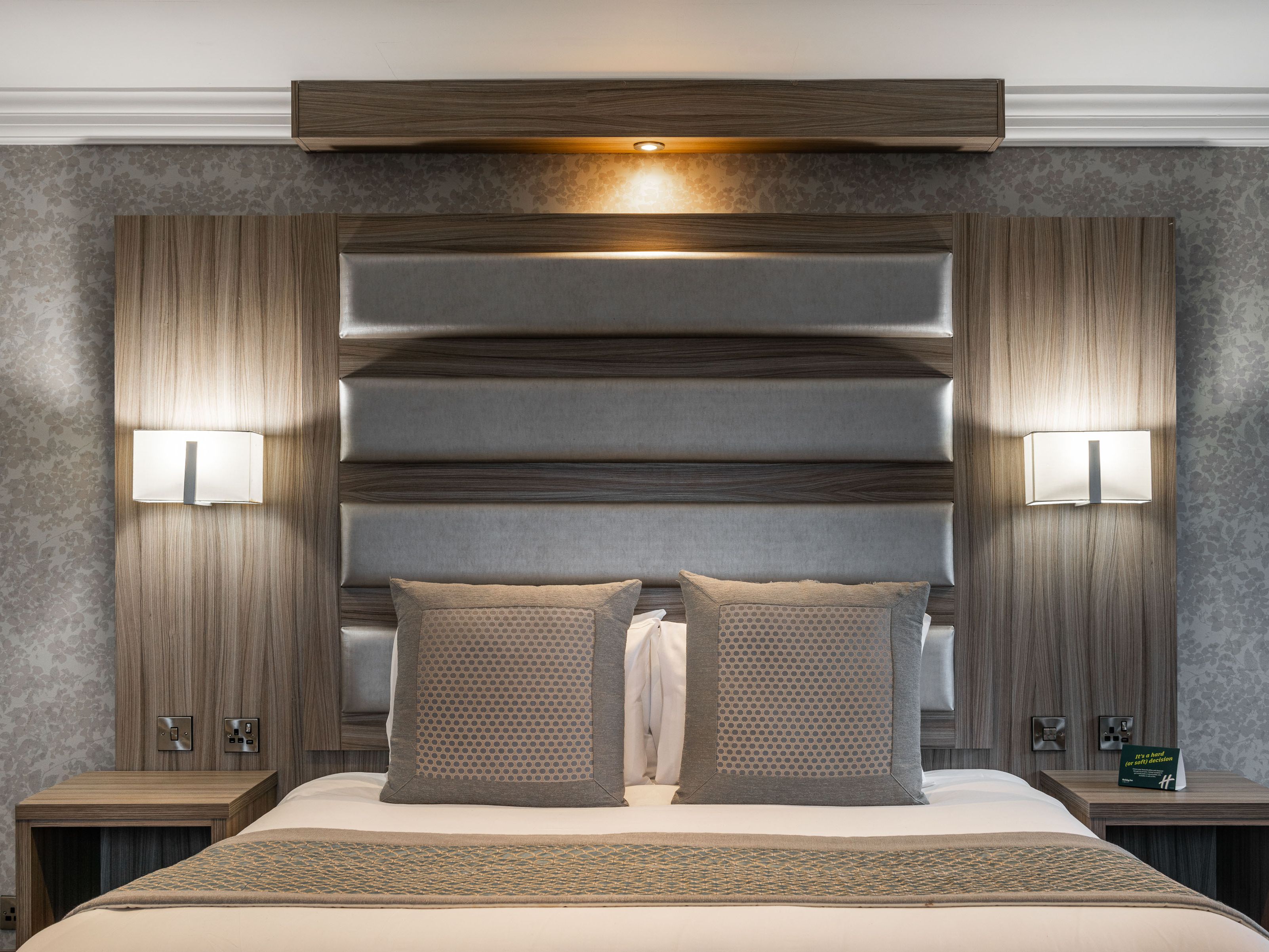 Modern hotel bedroom with a large bed, two square pillows, padded headboard, and built-in wall sconces, featuring wood accents and patterned wallpaper.