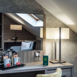 A cozy hotel room corner with a desk, modern lamp, tea and coffee set, mirror reflecting a gray armchair, and sloped ceiling with skylight window.