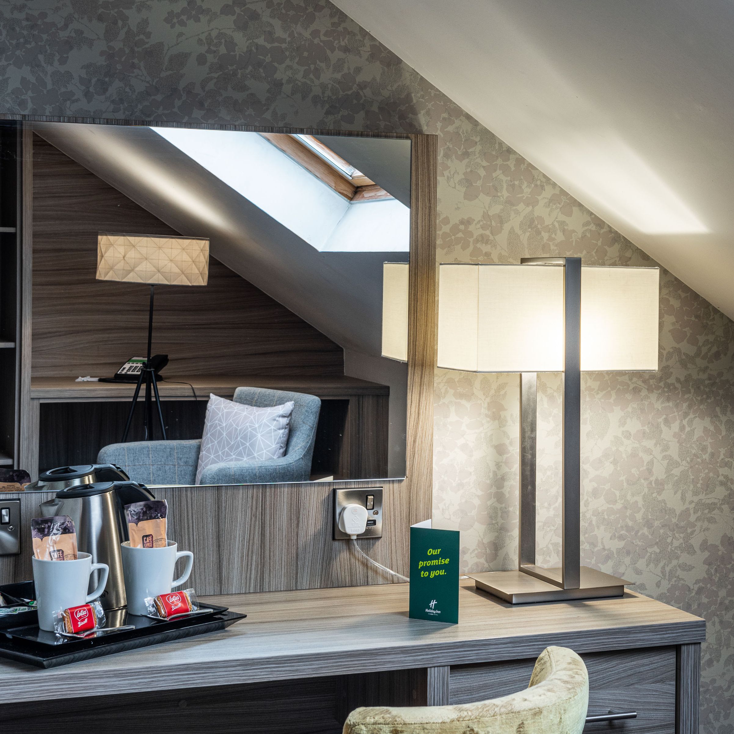 A cozy hotel room corner with a desk, modern lamp, tea and coffee set, mirror reflecting a gray armchair, and sloped ceiling with skylight window.