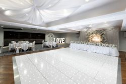 Elegant wedding reception hall decorated in white with draped ceiling fabric, floral arrangements, a long head table, illuminated dance floor, and large lit 'LOVE' letters in the background.