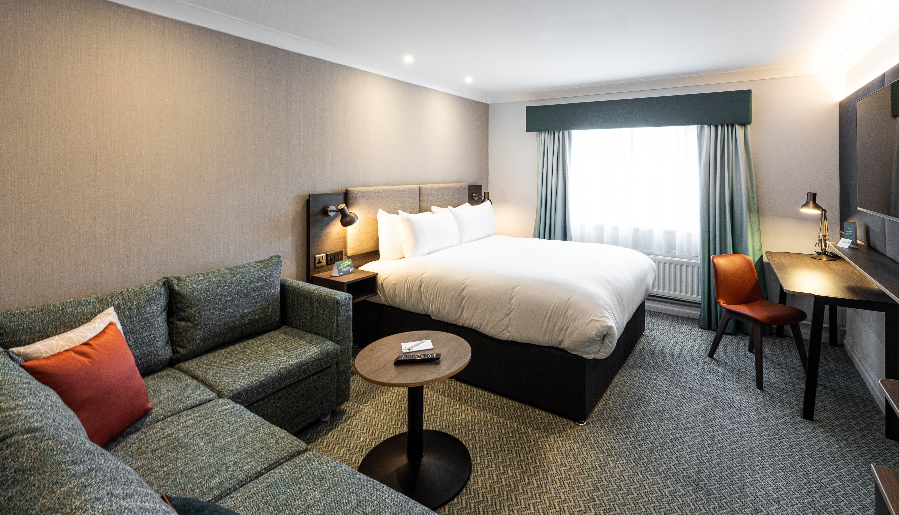 Modern hotel room with a large bed, green sectional sofa, round coffee table, desk with chair, and window with curtains.