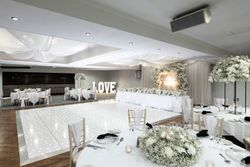 Elegant wedding reception hall decorated in white with floral arrangements, a twinkling dance floor, a long head table with white flowers, and large illuminated LOVE sign in the background.