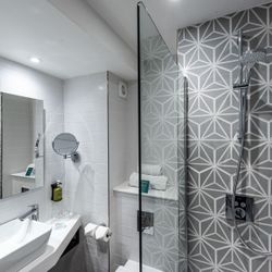 Modern bathroom with geometric patterned shower wall, glass shower partition, white sink, large mirror, and mounted toiletries.