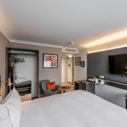 Modern hotel room with a large bed, flat-screen TV, work desk, sitting area with a chair and table, open closet with hangers and towels, and contemporary decor.