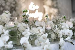Elegant table centerpiece with white flowers, including roses and orchids, and glass vases with floating candles at a wedding or formal event.