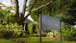 A large sign reading 'Welcome to Smokies Park' situated in a landscaped garden with green foliage, trees, and a statue in the background.