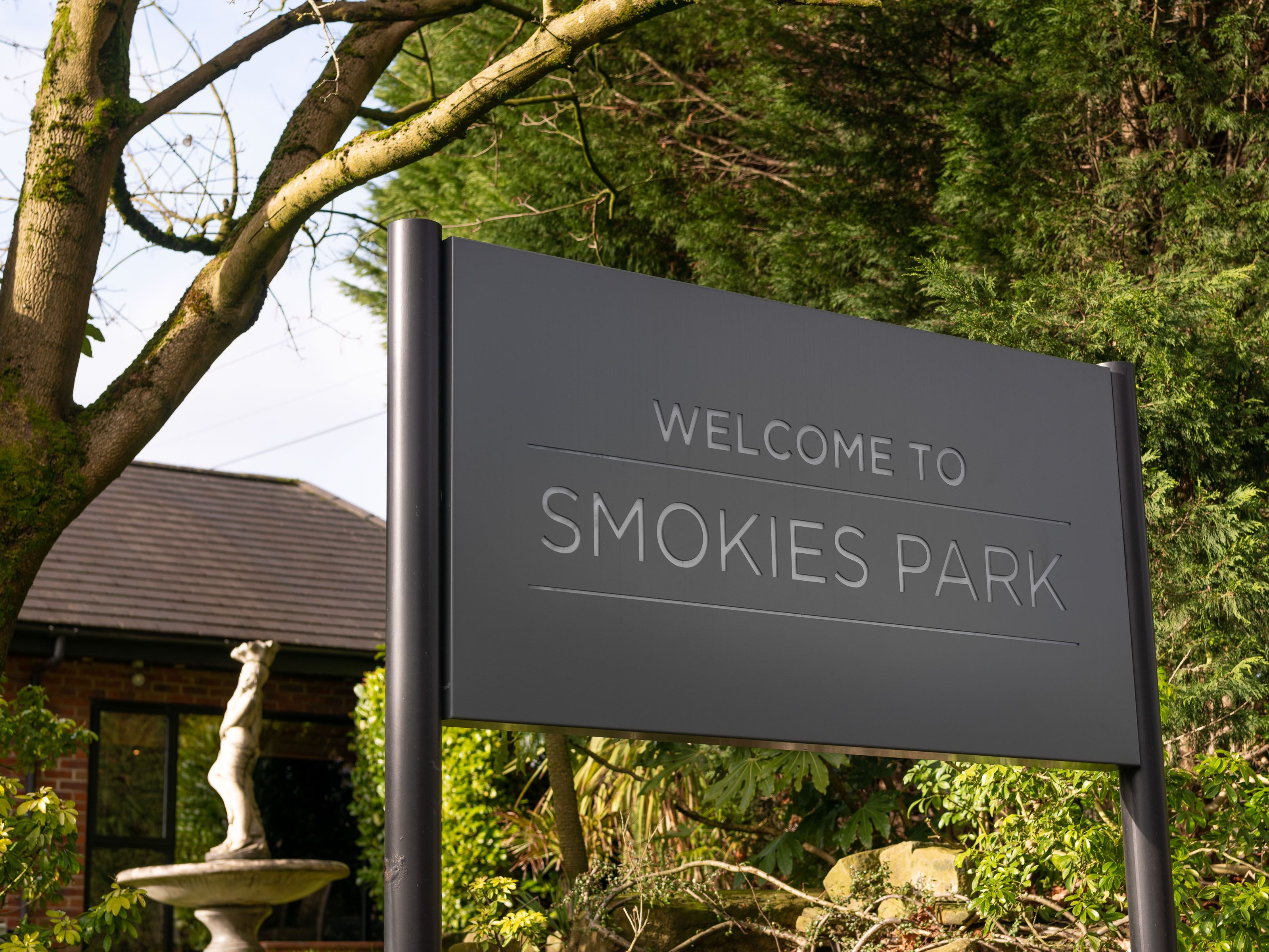 A sign that says 'Welcome to Smokies Park' surrounded by trees and greenery, with part of a building and a statue fountain in the background.
