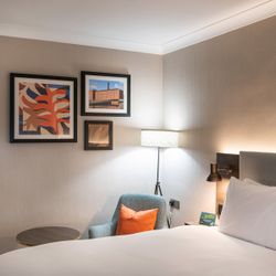 Modern hotel room with neatly made bed, cushioned chair with orange pillow, round side table, wall art, floor lamp, and bedside reading light.