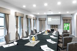 A modern conference room set up for a meeting with a large table, chairs, bottled drinks, glasses, notepads, pens, and a projection screen displaying the Holiday Inn logo.