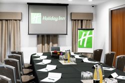 Conference room setup with a large table, chairs, notepads, pens, water bottles, glasses, a projector screen displaying the Holiday Inn logo, and a flip chart with the same logo.
