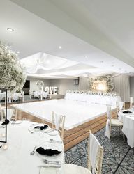 Elegant wedding reception hall decorated in white with floral centerpieces, round tables, a lighted dance floor, and a backdrop with large 'LOVE' letters.