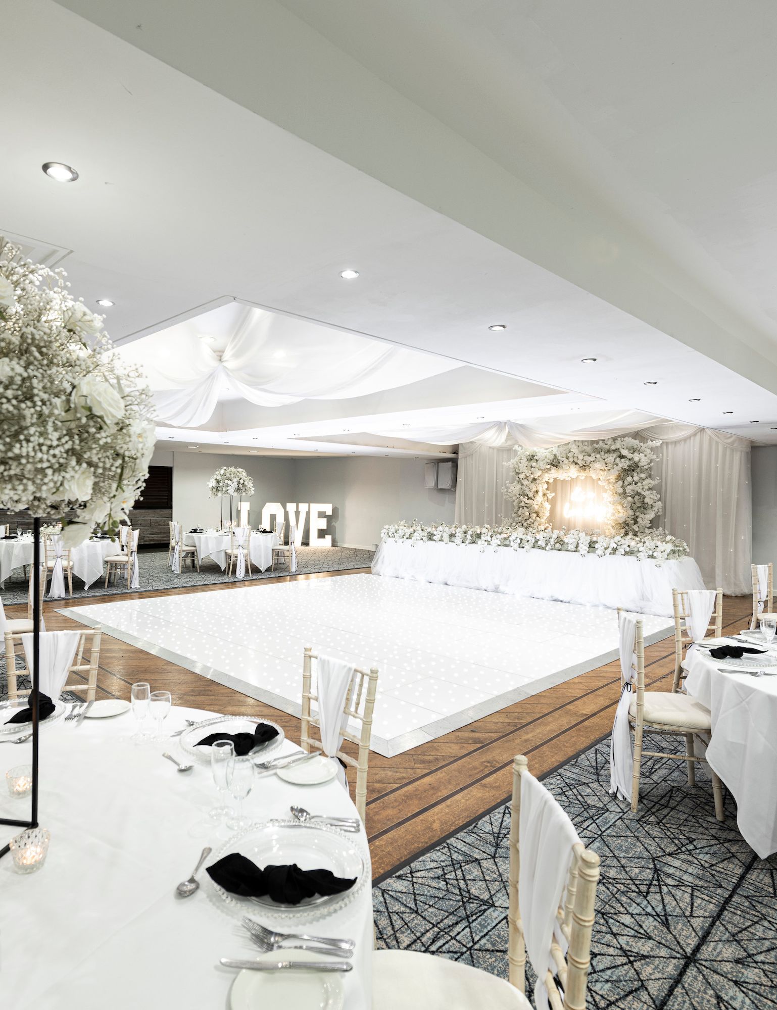 Elegant wedding reception hall decorated in white with floral centerpieces, round tables, a lighted dance floor, and a backdrop with large 'LOVE' letters.