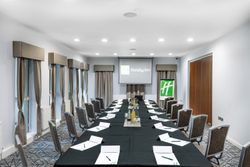 A modern conference room at Holiday Inn set up for a meeting with a large U-shaped table, chairs, notepads, pens, beverages, and a projector screen displaying the Holiday Inn logo.