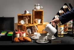 A coffee and pastry station with cups, coffee beans, pastries, yogurt parfaits, tea bags, syrup bottles, and a person pouring coffee from a white pot into a cup.