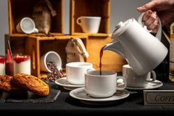A hand pours coffee from a white teapot into a white cup, with pastries, yogurt desserts, coffee beans, cups, and rustic wooden crates in the background.