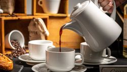 A hand pours coffee from a white teapot into a white cup, with pastries, yogurt desserts, coffee beans, cups, and rustic wooden crates in the background.