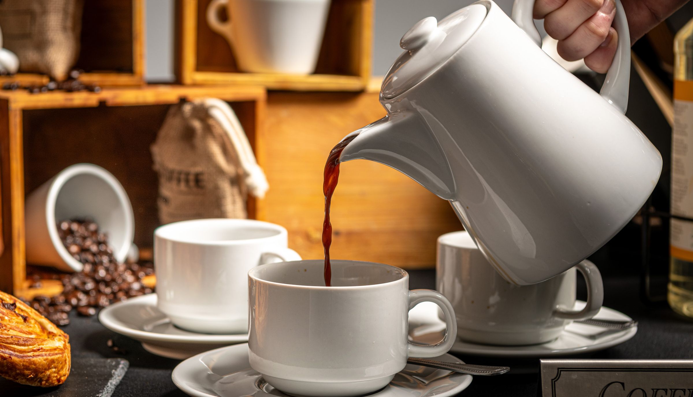 A hand pours coffee from a white teapot into a white cup, with pastries, yogurt desserts, coffee beans, cups, and rustic wooden crates in the background.
