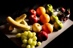 A variety of fresh fruits including bananas, apples, oranges, green apples, grapes, nectarines, and a jar filled with strawberries, arranged on a rectangular board against a dark background.