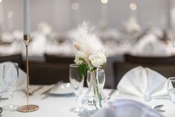Elegant table setting with white folded napkins, glassware, a gold candle holder, and a small vase containing white flowers and decorative feathers.