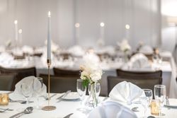 Elegant table setting with white linens, glassware, a tall white candle, and a small floral arrangement, prepared for a formal event or wedding reception.