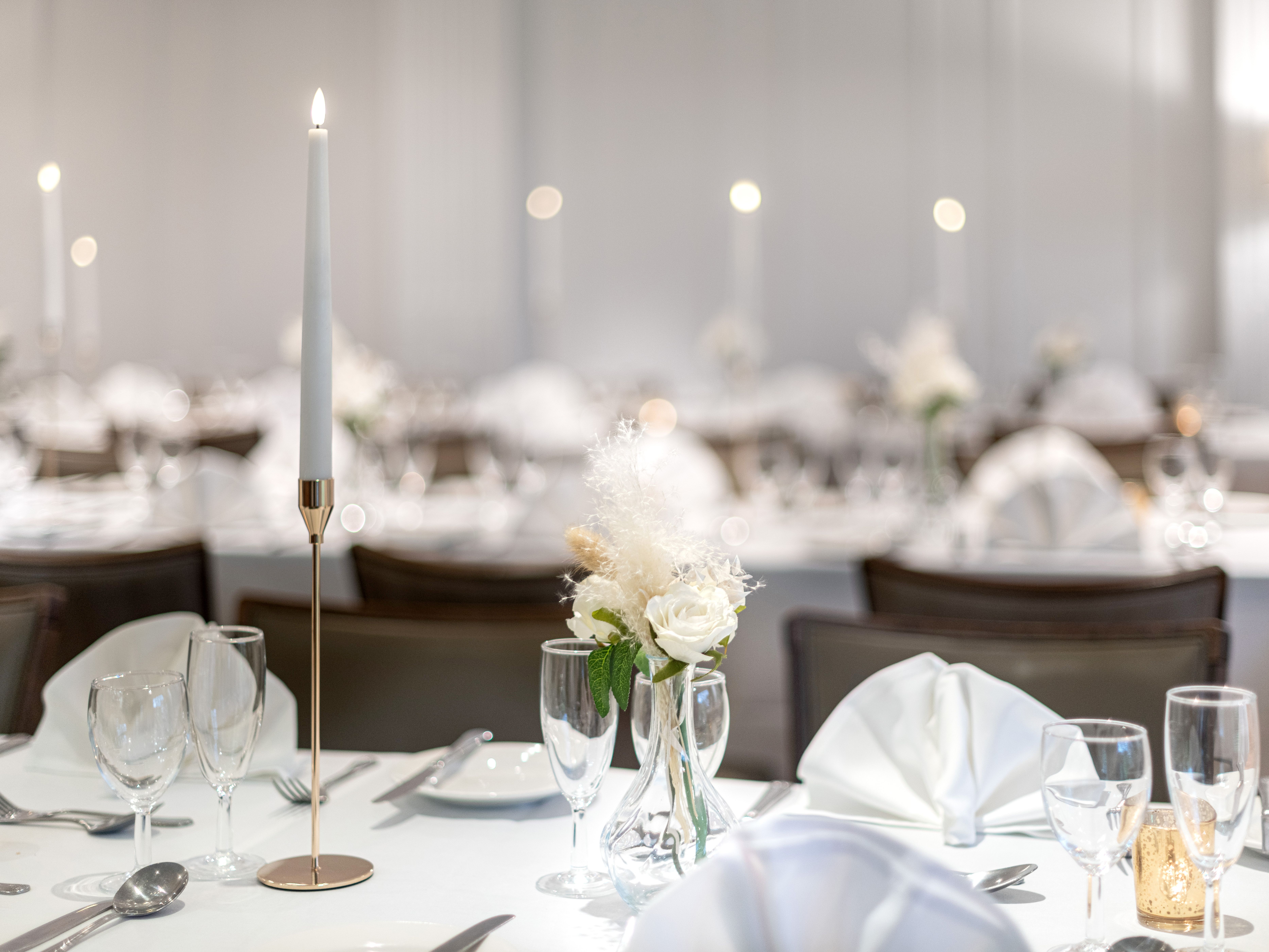 Elegant table setting with white linens, glassware, a tall white candle, and a small floral arrangement, prepared for a formal event or wedding reception.