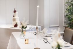 Elegant wedding table setting with white tablecloth, glassware, a candle, a small vase of flowers, and a blurred view of a tiered wedding cake decorated with flowers in the background.